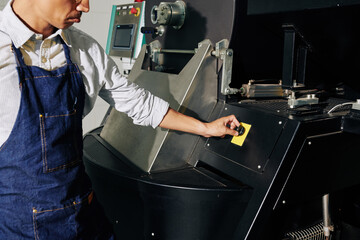 Cropped image of worker in denim apron turning controller on coffee roaster to ser rotating speed