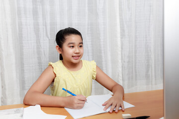 Naklejka premium Asian young little girl using pencil to do homework by herself. Student kid writing homework book. Girl use computer to study at home. Education homeschool concept.