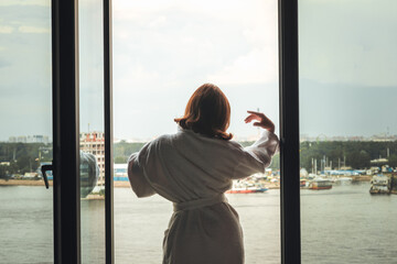 Obraz premium Back view of beautiful young woman in bath terry robe with cup of coffee standing and enjoying near open panoramic window of apartments opposite the lake.