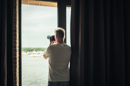 Back view of middle-aged gray-haired man photographer in a  taking picture using camera standing near open window of apartment or hotel room.