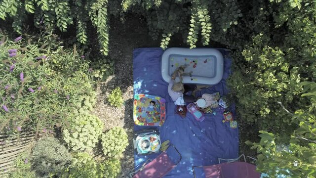 Blond mother helping her little child to bath in inflatable swimming pool in the lush summer garden. Baby sits in small square pool with color balls. Ascending aerial top shot.