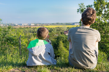 Dwaj chłopcy siedzą na skarpie wzgórza, odwróceni plecami, patrzą na horyzont - obrazy, fototapety, plakaty