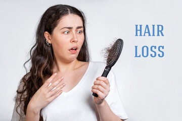 Hair loss. A terrified brunette woman holds a comb in her hands, with a bunch of hair that has fallen out. White background. Concept of baldness and hair care