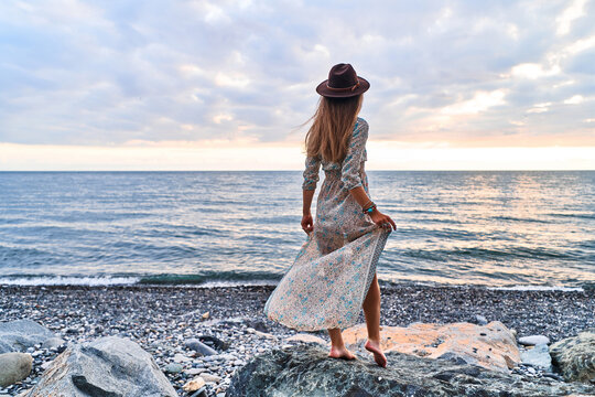 Boho chic woman in long dress and felt hat standing back on stone by the sea