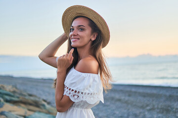 Portrait of beautiful young happy smiling romantic woman in a summer white dress with bare shoulders and a straw hat by the seashore at summertime