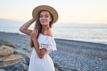 Portrait of beautiful young happy smiling romantic woman in a summer white dress with bare shoulders and a straw hat by the seashore at summertime