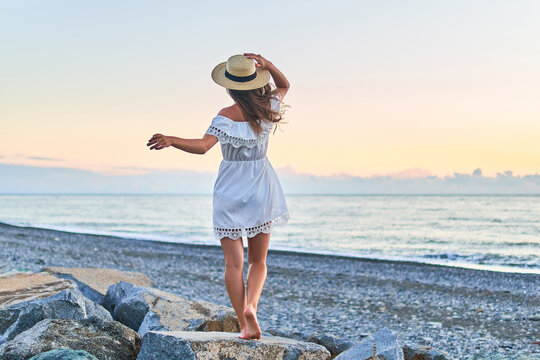 Young free romantic woman in a short summer white dress with bare shoulders and a straw hat standing on a stone by seashore at summertime