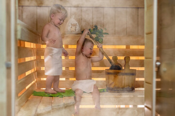 Kids are steaming in the Russian bath. Two kids with a bucket and a basin in the sauna. Twin boys relax in the bath. Сute children in the sauna on a wooden shelf. Steam and heat in the Russian sauna