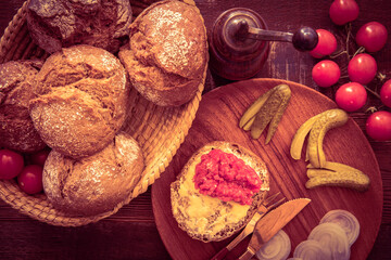 Bread rolls in basket with buttered roll minced meat onion cucumber on wooden plate vine tomato and pepper grinder on wooden table