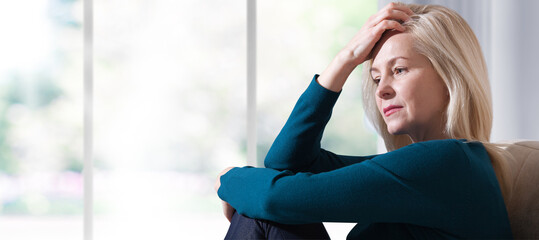 Woman suffering from stress or a headache grimacing in pain. Sad depressed woman at home sitting on the couch, looking down and touching her forehead.