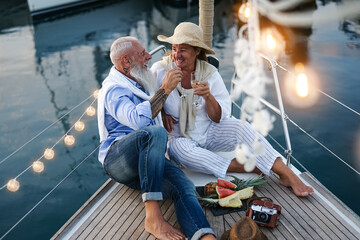 Senior couple toasting champagne and eating fruits on sailboat vacation - Happy elderly people...