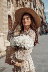 Young fashionable happy smiling woman wearing straw hat, floral print dress, holding wicker bag...