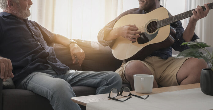 Caucasian old father and son happy spending time together; talking, play guitar, sing a song in cozy living room warmth house on holiday with coffee cup and glasses on the table.