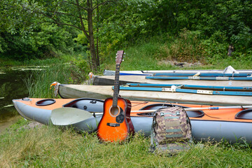 A guitar and a backpack stand against the backdrop of multi-colored kayaks on the river bank. Vacation, hiking, travel