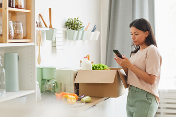 Waist up portrait of modern mixed-race woman holding smartphone while standing by box of food in kitchen, mood delivery service and mobile app concept