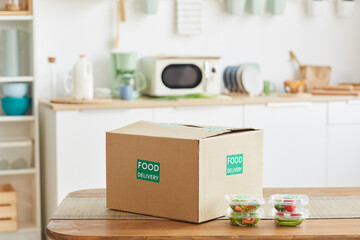 Background image of cardboard box with food standing on wooden table in white kitchen interior, food delivery service, copy space