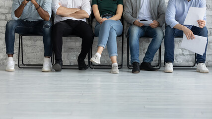 Fototapeta na wymiar Cropped image legs of diverse candidates waiting for job interview, applicants business people sitting in row in queue, holding resume documents, human resources and recruitment concept