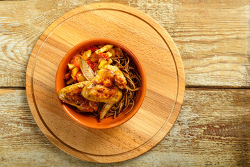 Brown cup with soba noodles with vegetables and chicken wings on a wooden stand.