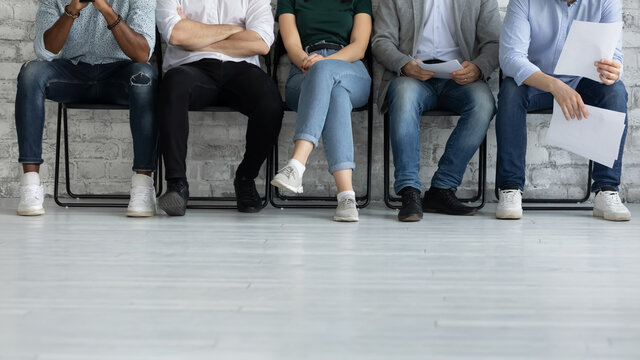 Cropped image legs of diverse candidates waiting for job interview, applicants business people sitting in row in queue, holding resume documents, human resources and recruitment concept