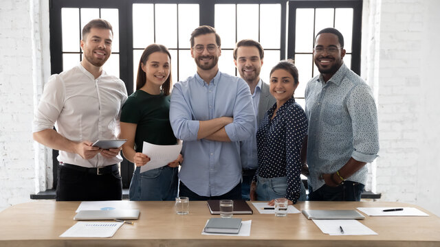 Corporate photo smiling diverse employees with confident executive wearing glasses standing in modern office room, looking at camera, successful startup founder with team, staff members
