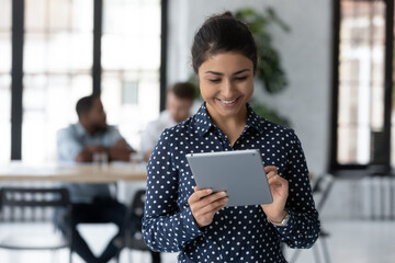Close up smiling Indian businesswoman holding digital tablet, standing in modern office room, employee working online, using apps, writing or reading email, chatting in social network during break