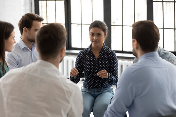 Confident Indian psychologist coach speaking at meeting group training, supporting diverse people sitting in circle at therapy session, mentor training staff, team building activity in office