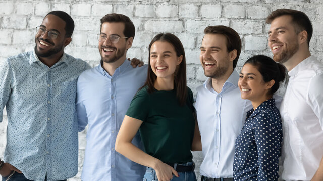 Smiling motivated successful diverse employees team standing in modern office close up, looking to aside, posing for corporate photo, hugging, confident happy business people staff department