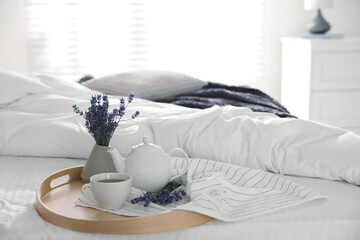 Wooden tray with cup of fresh hot tea and beautiful lavender flowers on bed, space for text. Tasty breakfast