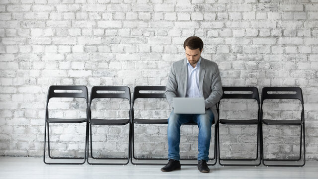 Confident serious businessman candidate applicant seeker using laptop, waiting, preparing for job interview, sitting on chair in empty office hallway, recruitment and employment concept