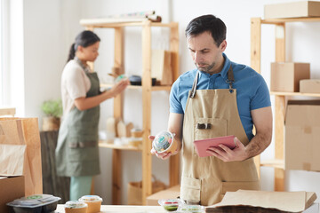 Waist up portrait of mature worker wearing apron packaging orders while standing by wooden table, food delivery service, copy space