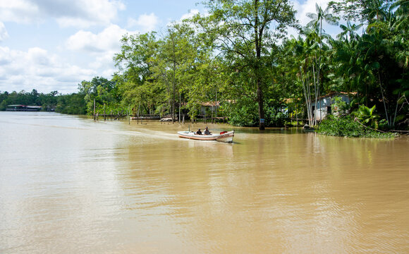 Duas pessoas andando de barco de madeira a motor em rio na margem da floresta amaz&ocirc;nica, no Par&aacute;. Casas ribeirinhas na margem da &aacute;gua.