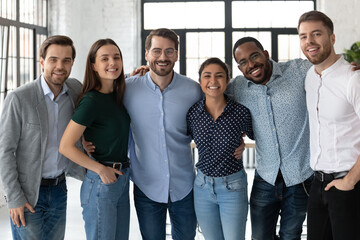 Smiling diverse business people, successful team, staff members hugging, standing in modern office, looking at camera, happy overjoyed employees colleagues posing for corporate photo - obrazy, fototapety, plakaty