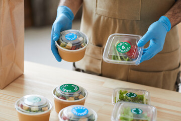 Closeup of unrecognizable worker wearing protective gloves safely packaging orders at wooden table in food delivery service, copy space