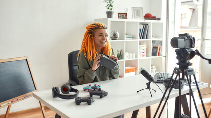 Female technology blogger with dreadlocks holding vr glasses while recording video review of new gadgets using microphone at home