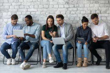 Diverse young candidates using gadgets while waiting for job interview or exam, sitting in row in...