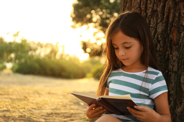 Cute little girl reading book near tree in park