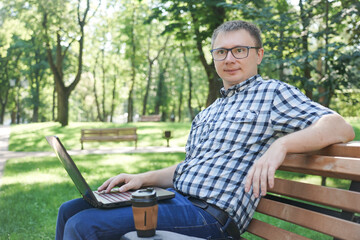 Fototapeta premium A guy with a laptop sits on a park bench. A cup of coffee stands near the laptop.