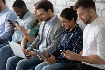 Close up smiling diverse business people candidates waiting for job interview, sitting in row in queue, using electronic devices, phones, laptop, human resources and employment concept