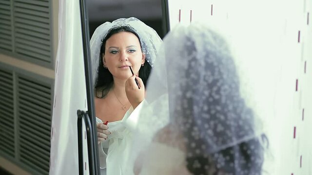 A Jewish bride in a dress and a veil in front of a mirror applies makeup before the hupa ceremony, reflected in the mirror