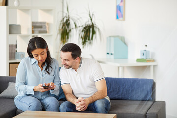 Portrait of young mixed-race woman showing smartphone to male colleague while sitting on couch in office interior, copy space