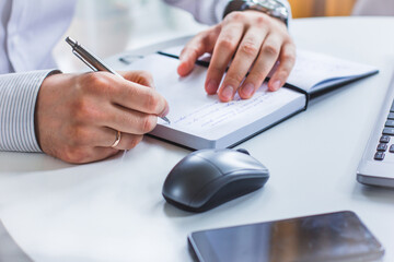 An image of the hands of a man sitting at a table and writing in a Notepad with a ballpoint pen. Next to it is a laptop, a computer mouse, and a cell phone
