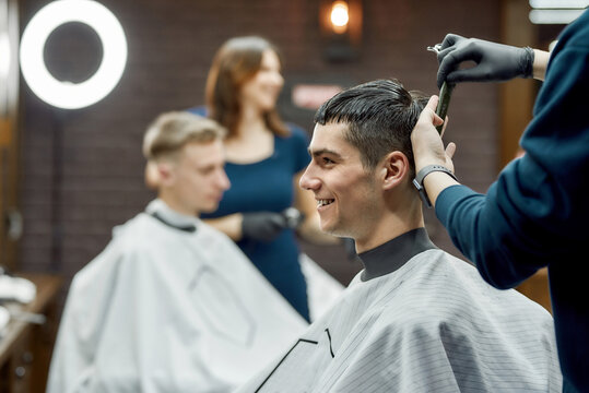 Side view of a young cheerful handsome guy visiting barbershop or hair salon, professional barber making new trendy haircut for client