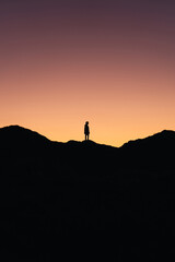 Vertical photo of a person standing in the middle of the mountain with the colors of the sunset in the background
