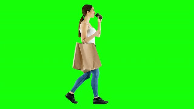 Caucasian woman walks with shopping bags, drinking coffee on green screen