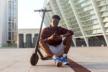 afro american guy sits with an electric scooter and uses a smartphone in the city, a student with eco transport holds a phone - Powered by Adobe