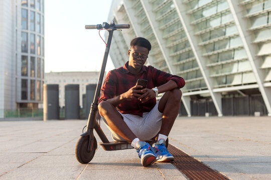 afro american guy sits with an electric scooter and uses a smartphone in the city, a student with eco transport holds a phone