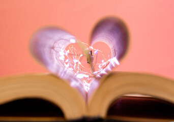 Open book with heart shaped sheets decorated with bright garlands of lights and highlights in the background, romantic love symbol, macro shot. Pink background