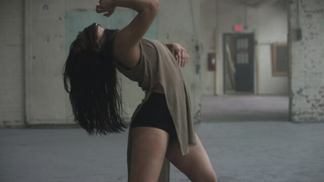 Beautiful dancer whips her hair leaning back, dancing passionately surrounded by mist in industrial warehouse