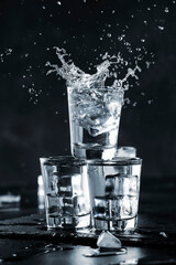 Vodka splash with spray from shot glasses on black stone background, iced strong drink in misted glass