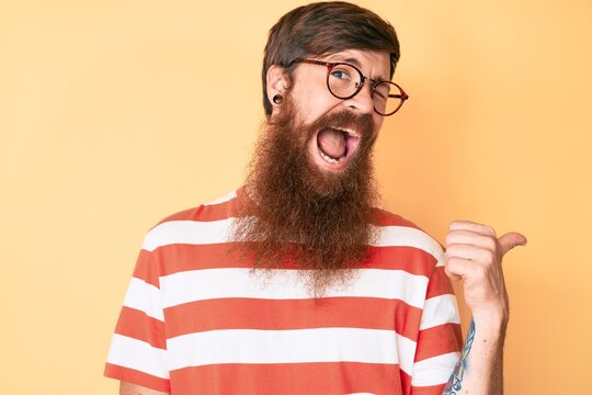 Handsome young red head man with long beard wearing casual clothes and glasses pointing thumb up to the side smiling happy with open mouth
